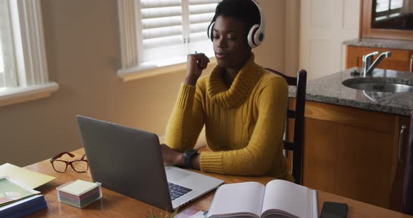 African american woman wearing headphones using laptop while working from home alt