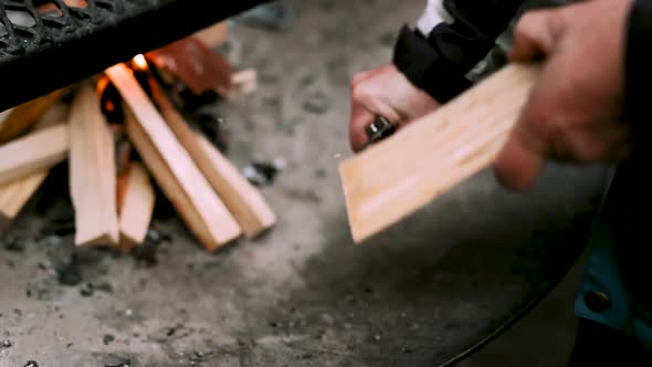 man cutting wood shavings of a log besides campfire alt