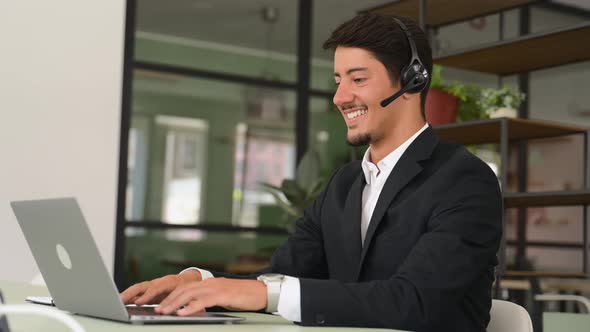 Friendly and Smiling Hispanic Man Wearing Wireless Headset Looks at the Camera Sitting at the Desk
