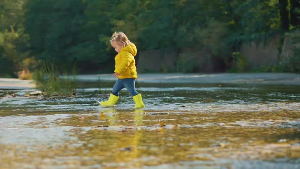 A cute baby girl in yellow rubber boots walking in the water, on the river. Slow motion. alt