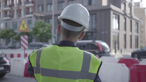 Little Boy Wearing Business Suit and Safety Equipment and Constructor Helmet Standing on a Busy Road alt