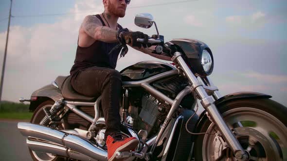 Stylish Biker with Tattoos Rides a Motorcycle on a Country Road at Sunset