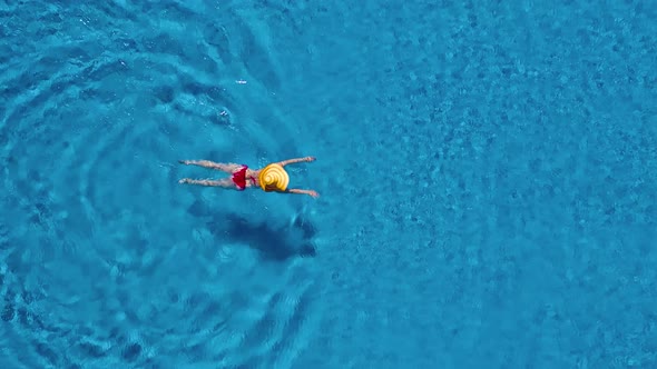View From the Top As a Woman in a Red Swimsuit and a Big Yellow Hat Swims in the Pool alt