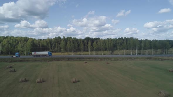 The Drone Flies Over the Field with Bales to the Forest Where There is a Track Along Which Trucks