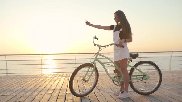 Young Attractive Woman Doing Selfie on a Bicycle at Sunrise or Sunset Near the Sea alt