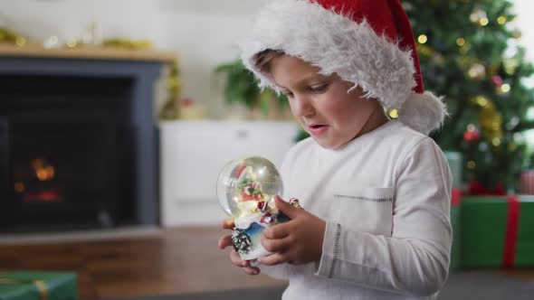 Surprised caucasian boy wearing santa hat playing with snow globe alt
