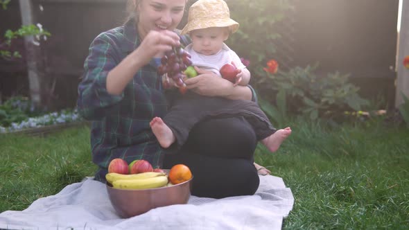 Happy Young Cheerful Mother Holding Baby Eating Fruits On Green Grass