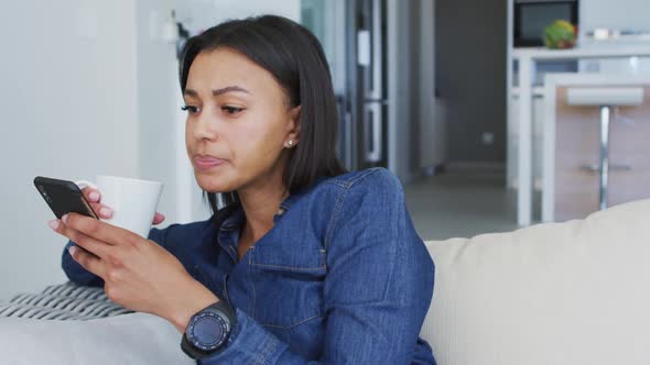 Mixed race woman sitting on couch using smartphone drinking cup of coffee alt