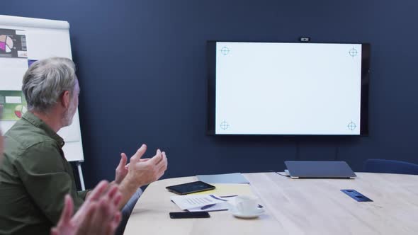 Diverse business colleagues sitting looking at screen and clapping in office meeting alt
