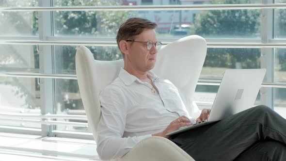 Mature Businessman Sitting on a White Chair in a Glass Office and Working on a Laptop