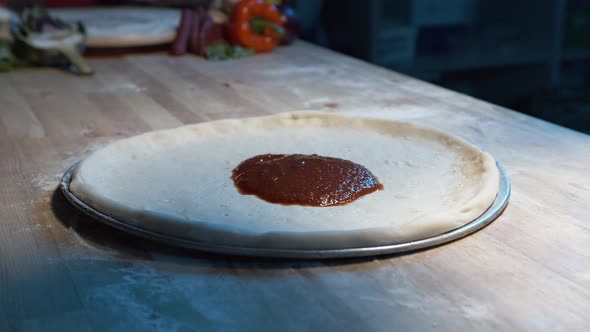 pizza dough on a steel tray on wooden counter while an expereinced chef put red sauce on it with a a alt