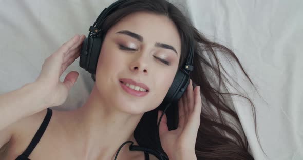Happy Girl Listening to Music on Bed Flirts at Camera Twists Headphones' Wire alt