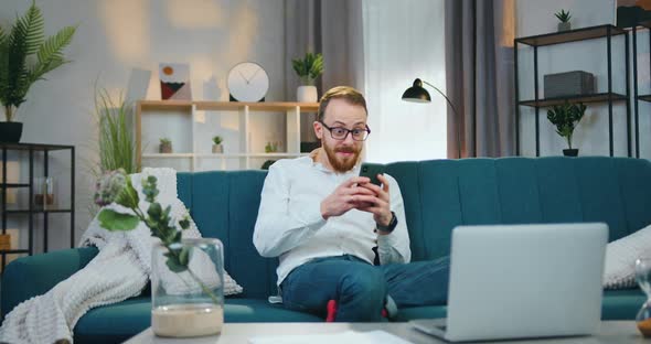 Man in Glasses Rejoycing when Get Good News on Smartphone and Raising His Hands from Happiness alt