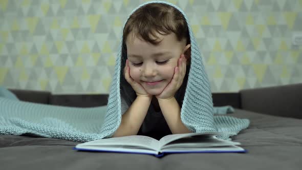 a Boy Reads an Interesting Book Lying in Bed Under a Blanket