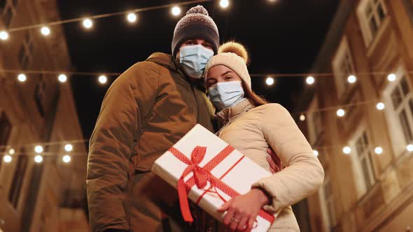 Couple in Protective Medical Face Masks Celebrating Xmas Eve Together at Bright Garland Decoration