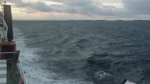 Man Standing On Ship Deck Leaning On Railing Looking Out Over Stormy Ocean Surface Waves alt