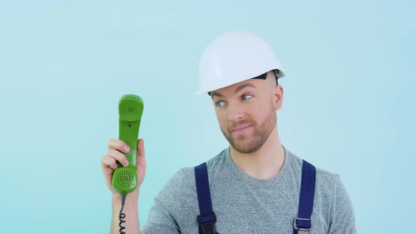 Repairman in Overalls and Helmet Holds an Old Phone in His Hands and Points to It alt