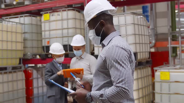African Manager in Safety Helmet with Clipboard Doing Inventory in Large Warehouse alt