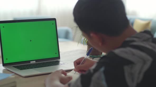 Boy Sitting At His Desk Learning Online On A Laptop With Green Mock-Up Screen alt