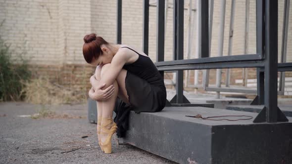 Sad Slim Caucasian Ballerina in Black Dress Sitting Outdoors Hugging Knees alt