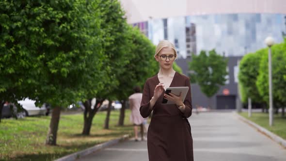 A Young Woman in a Business Suit and Formal Glasses Walks Down the Street and Scrolls the Tablet