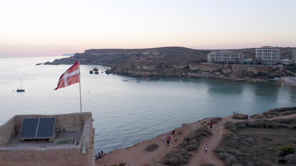 Flag of the Maltese order waving on a tower above a bay,sunset,aerial. alt