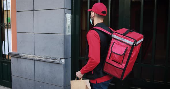 Young courier man with thermal backpack ringing the doorbell while wearing safety face mask alt