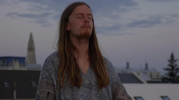 Young Man With Long Hair Meditating On City Roof alt