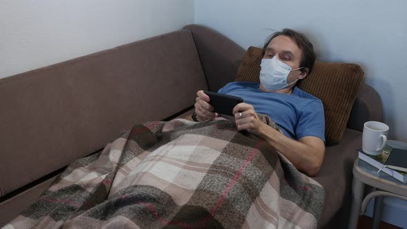 Sick Adult Man in Protective Mask Lying on a Bed and Using Smartphone 