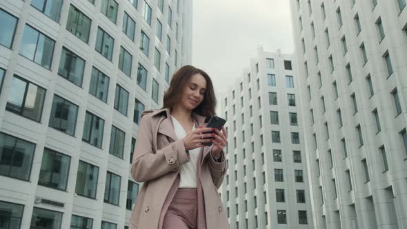 Cheerful young Caucasian woman standing in city street and texting on mobile phone with smile. alt