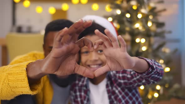 Portrait of African Father and Son Gesturing Heart with Hands Looking at Camera Near Christmas Tree alt