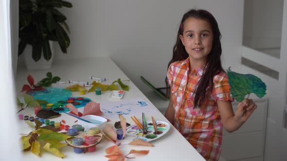 Closeup of Little Girl Sitting at Table Drawing the Leaves with Brush and Watercolours alt