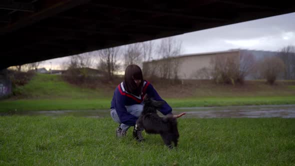 a Brunette in Jeans and a Blue Sweater Walks with Her Dog Near the River Under the Bridge. They Play alt