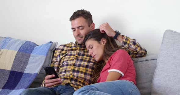 Cute man using mobile phone and woman relaxing on his shoulder in living room