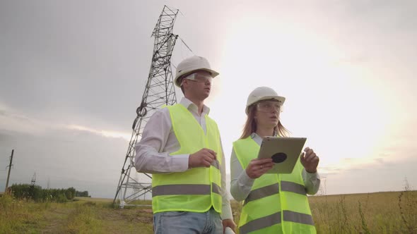 Two Engineers a Man and a Woman in Helmets with a Tablet of Engineer Walk on Field with Electricity alt