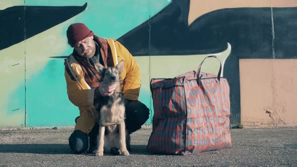A Tramp is Stroking His Dog with a Massive Bag Next to Them alt