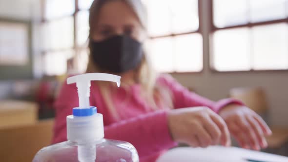 Girl wearing face mask sanitizing her hands while sitting on her desk at school alt