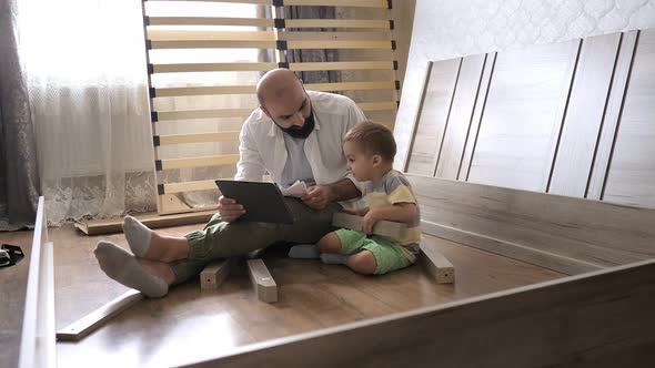Bearded Man with Son Sitting on the Floor Reading Instruction in the Tablet for Assembling Furniture