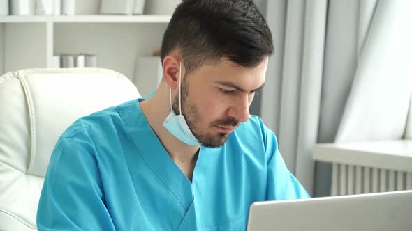 Male Doctor in Uniform and Mask Works at Laptop in Clinic