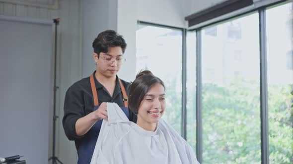 Asian professional hair stylist man puts protective cape apron to Caucasian beautiful woman in salon alt