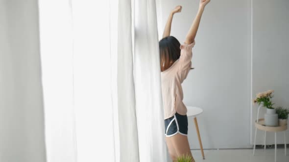 A young Asian woman opens the curtain and stretches Beside the window.