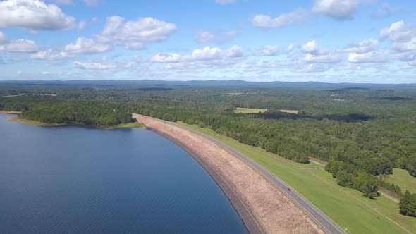 Cars Driving on a Lake Dam Surrounded by Forest and Blue Water and Distant Mountains alt