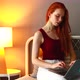 Redhaired Ginger Woman Sitting at Her Bedroom with Laptop on the Bed  Evening Lamp on