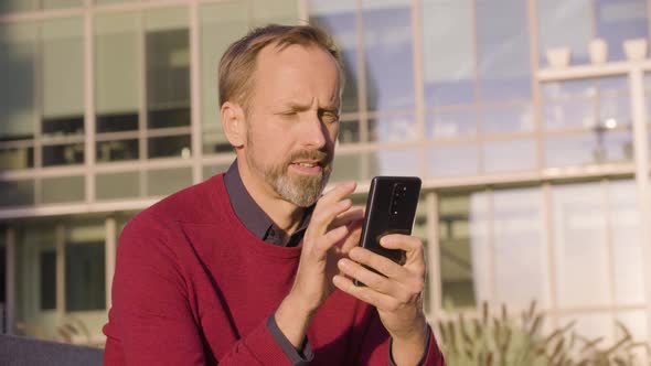 A Middleaged Handsome Caucasian Man Works on a Smartphone with a Smile As He Sits alt
