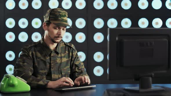 Man in Military Clothes Types on Keyboard Sits at Desk Next to Green Telephone