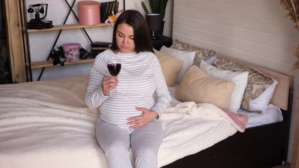 A Pregnant Woman with a Glass of Red Wine is Sitting on the Bed at Home alt
