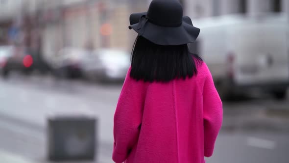 a Brunette in a Black Hat Walks Away Against the Background of a Blurry City Street Turns Around alt