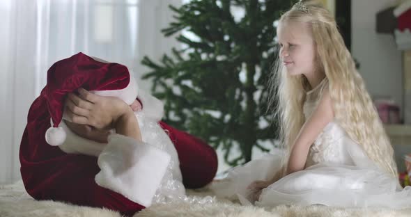 Positive Caucasian Girl with Blond Curly Hair Making Hush Gesture and Talking To Santa at Home alt