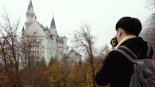 Male Asian Tourist with Backpack and Camera Standing and Enjoying View of Neuschwanstein Castle alt