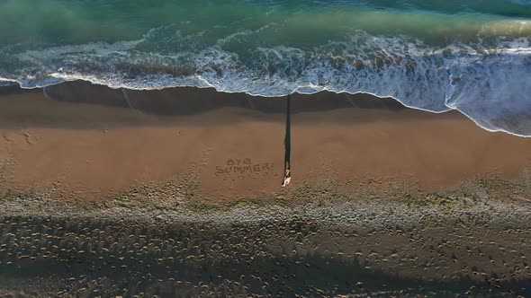 A woman writing the words "BYE SUMMER" in the sand alt